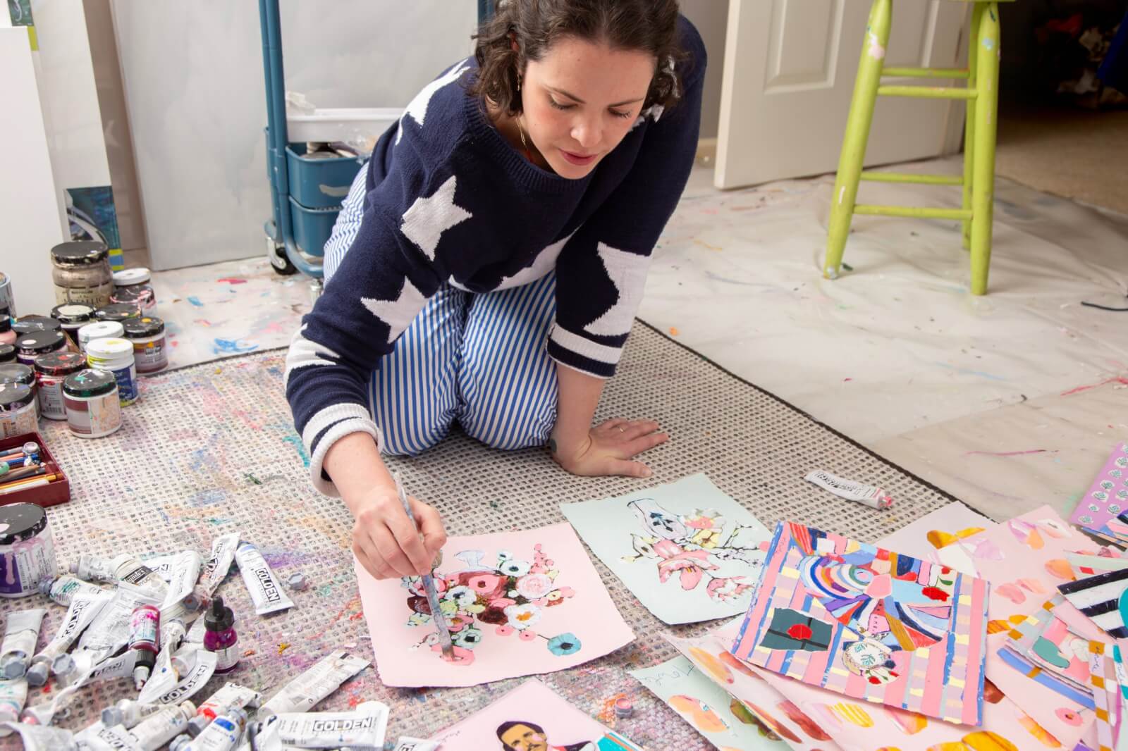 Meet Memphis Artist Heather Howle - 1 A woman kneels on the floor painting a floral picture on pink paper, surrounded by art supplies, paint tubes, colorful finished artworks, and sketches.