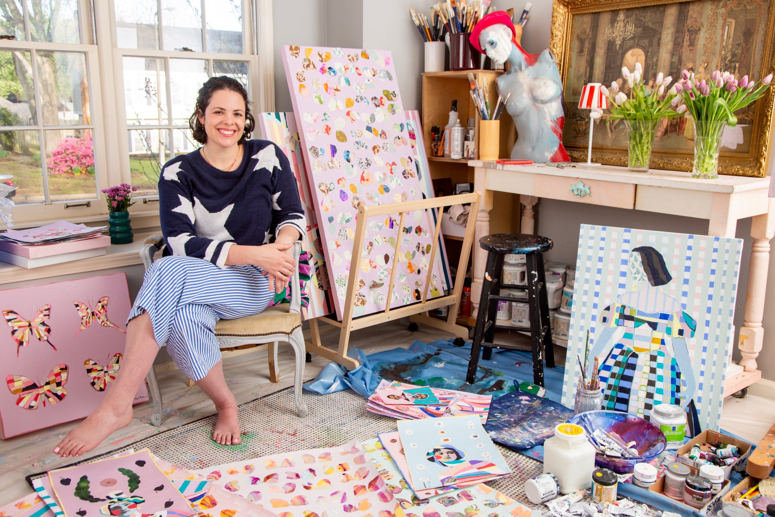 Meet Memphis Artist Heather Howle - 3 Heather Howle sits barefoot in her art studio, surrounded by colorful paintings, art supplies, and a mannequin bust, as natural light streams through the window.