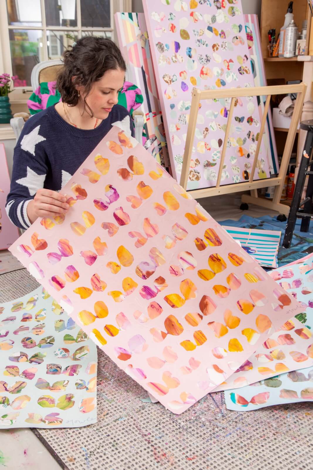 Meet Memphis Artist Heather Howle - 5 Heather Howle sits on the floor in her studio, holding a large sheet with colorful abstract patterns. More similar artworks and art supplies are visible around ehr.