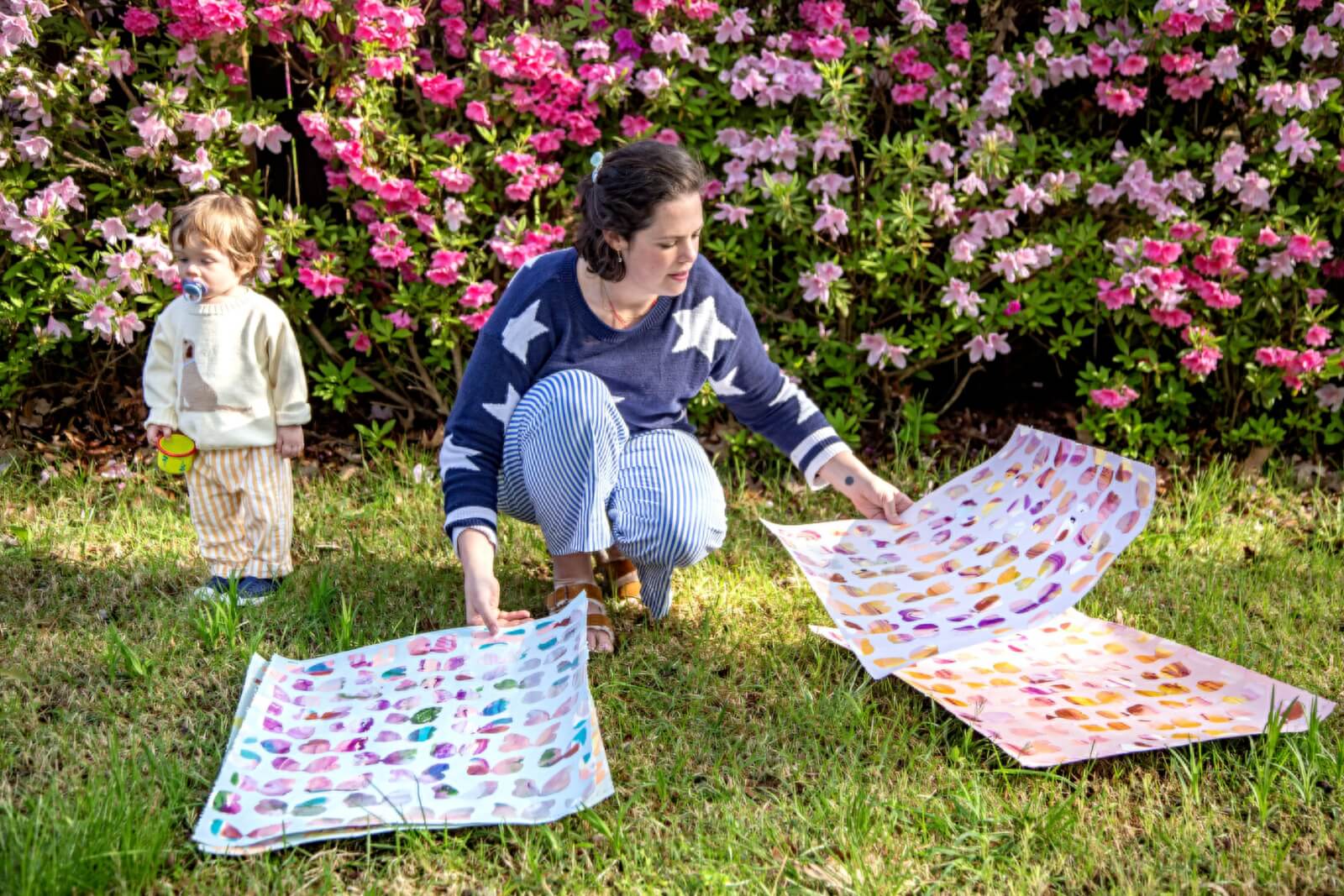Meet Memphis Artist Heather Howle - 4 Heather Howle kneels on grass arranging three colorful patterned sheets, while a toddler with a pacifier stands nearby. Pink flowers are in full bloom in the background.