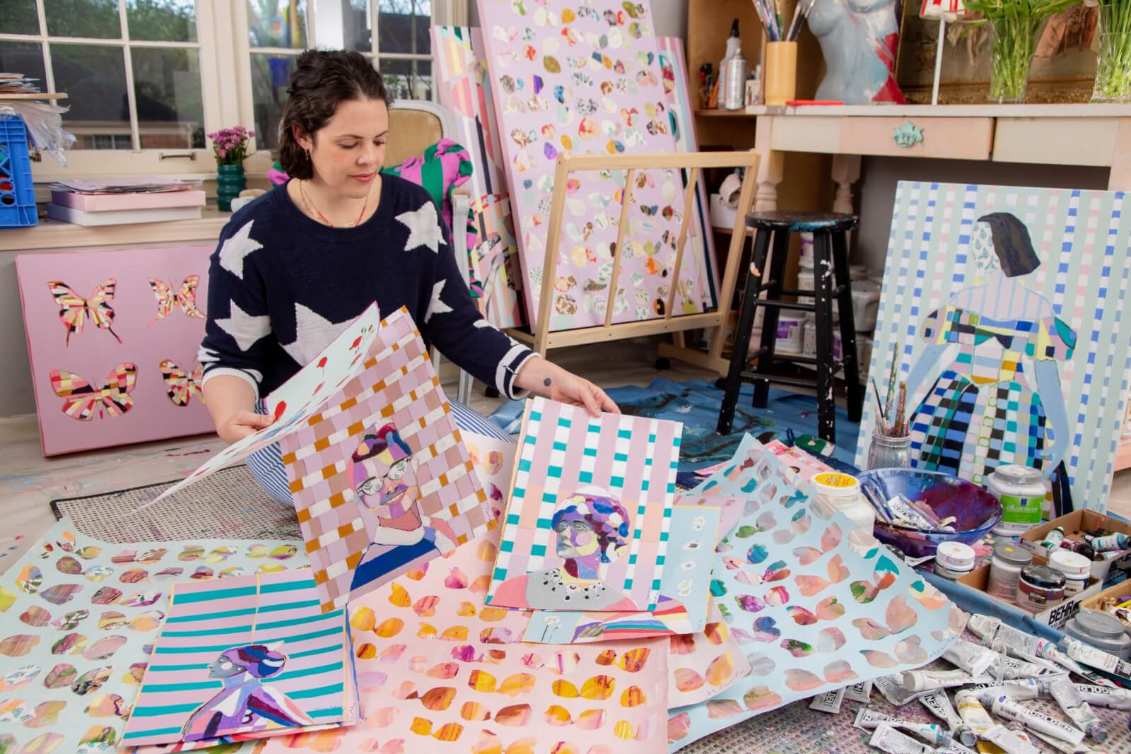 Meet Memphis Artist Heather Howle - 2 An artist sits on the floor in a studio, surrounded by colorful paintings and art supplies, examining a piece.