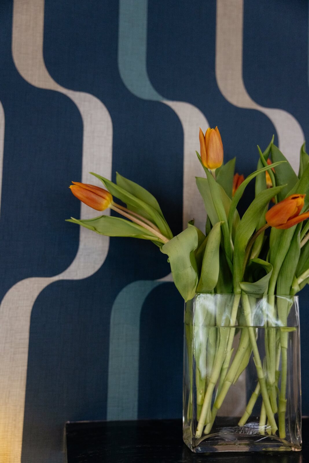 A clear glass vase with orange tulips sits in front of blue and beige geometric patterned wallpaper, evoking the vintage hotel charm of Hotel Pontotoc in Memphis.
