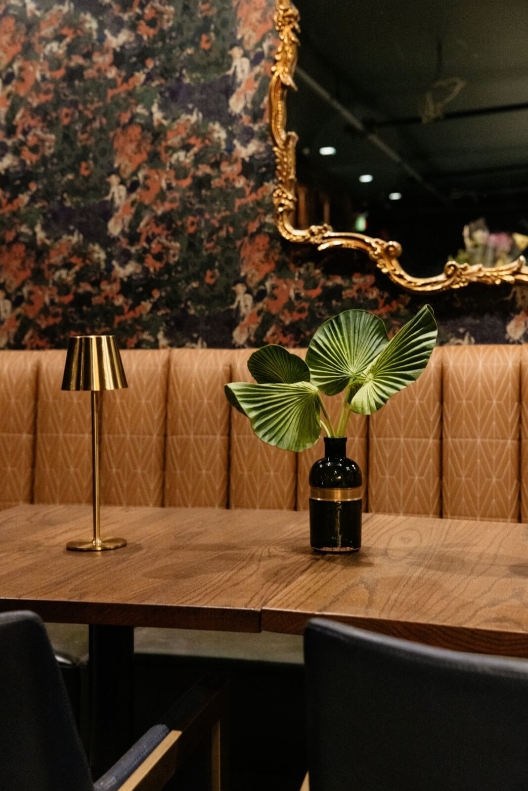 A small table with a brown leather booth, gold lamp, and green leaves in a black vase, set against a patterned wall and ornate gold-framed mirror at the vintage Hotel Pontotoc in Memphis.