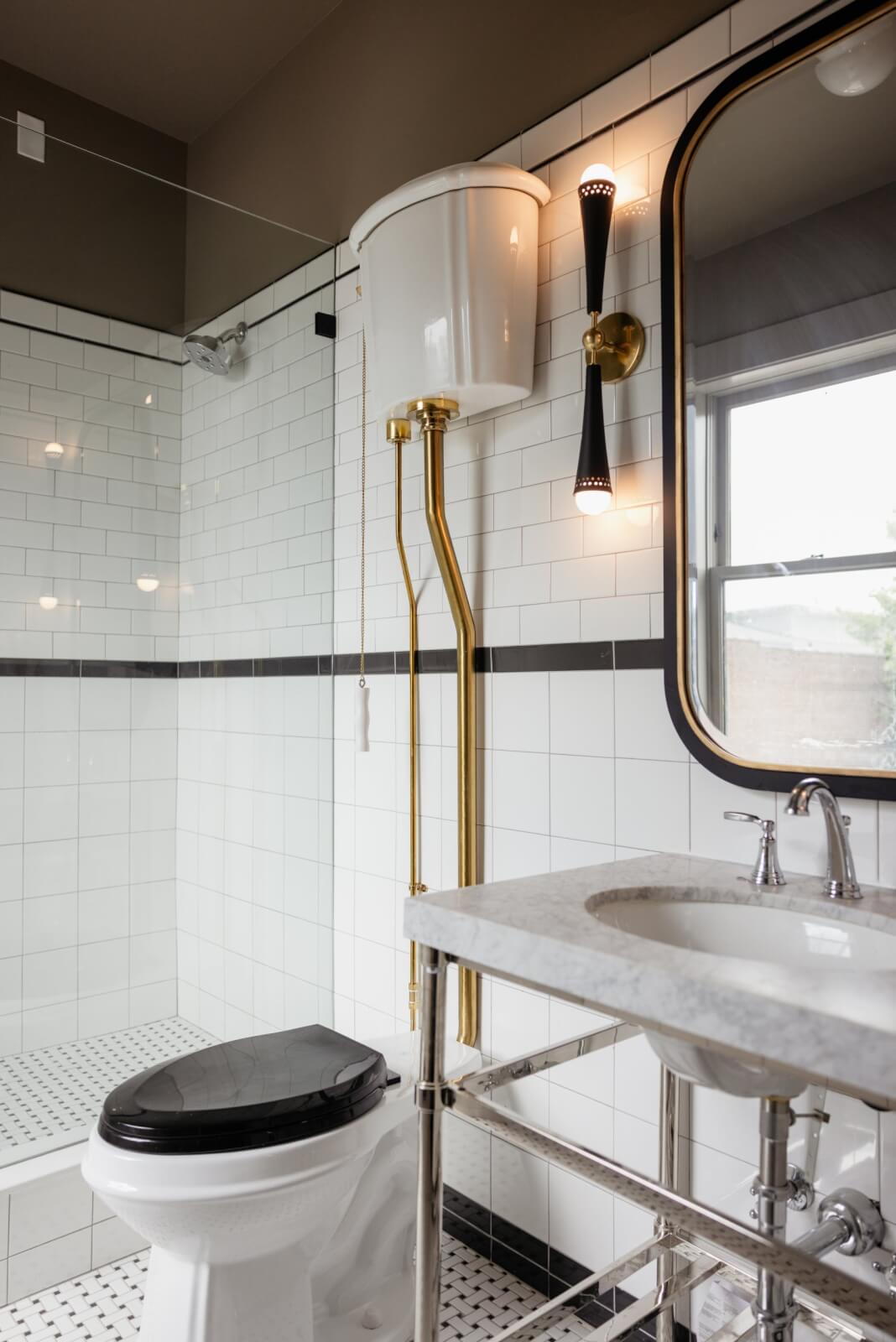 A bathroom in the vintage Hotel Pontotoc, Memphis, features white subway tile walls, a black toilet seat, a high-tank toilet with gold pipes, a marble sink, and a large mirror beside a wall sconce.
