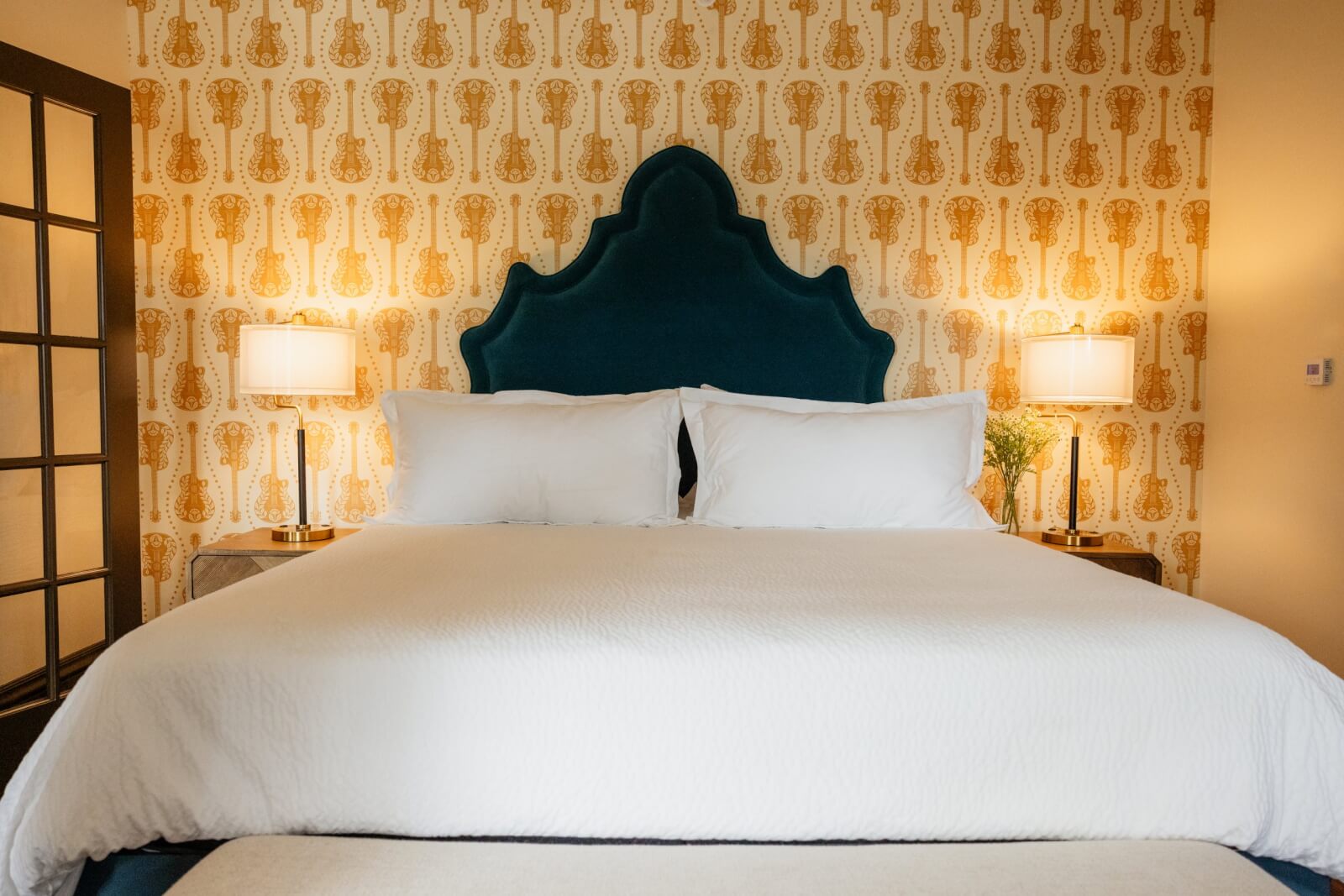 A neatly made bed with a dark blue, arched headboard, white bedding, and two lit lamps on side tables evokes the vintage hotel charm of Hotel Pontotoc in Memphis, set against stylish patterned wallpaper.