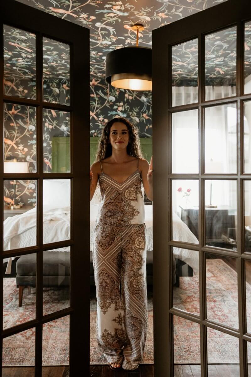 A woman in a patterned jumpsuit stands between open French doors in a stylish Hotel Pontotoc bedroom in Memphis, featuring floral wallpaper and soft natural light that evokes the charm of a vintage hotel.