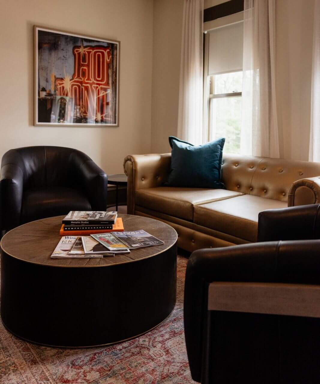 A cozy living room reminiscent of Hotel Pontotoc in Memphis, with a gold tufted sofa, two black armchairs, a round coffee table with books and magazines, large windows, and framed wall art adding to the vintage hotel charm.