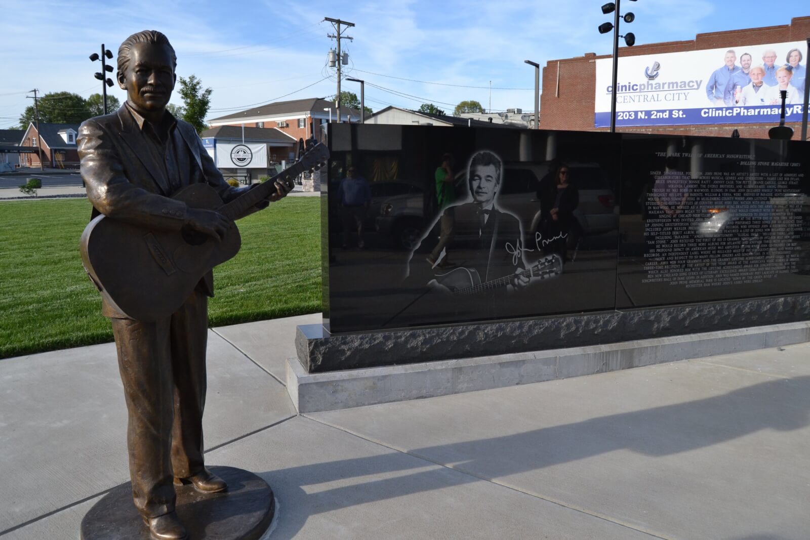 A bronze statue of a man playing guitar stands by a black granite monument engraved with his portrait and signature, creating a unique tribute in Western Kentucky near a grassy area and pharmacy building.
