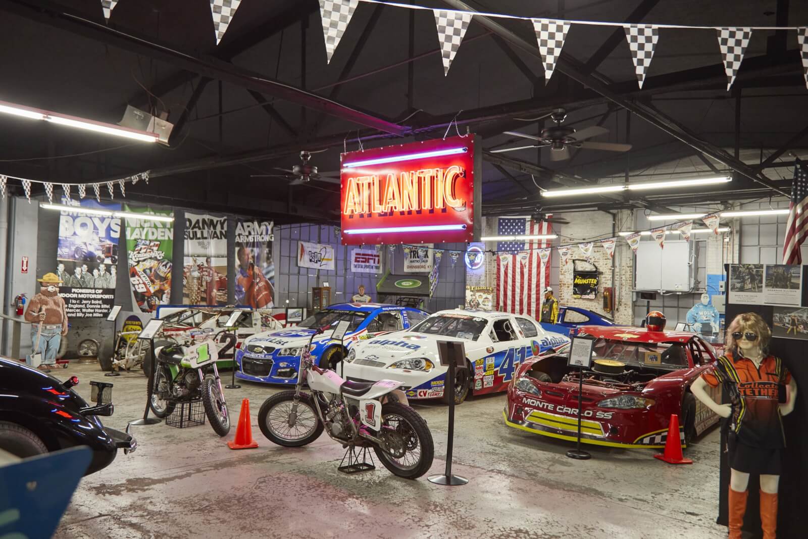 Indoor display of race cars and motorcycles with a red "ATLANTIC" neon sign overhead; posters and flags decorate the background.