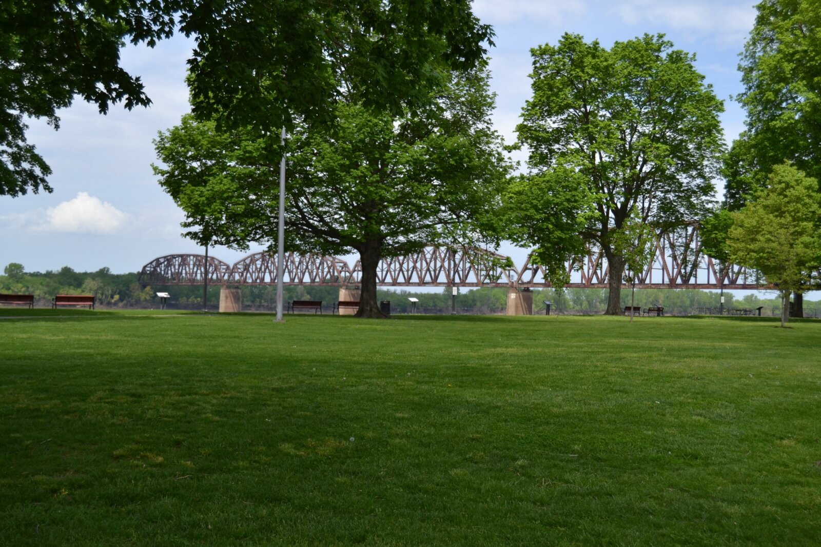 A large grassy park with scattered trees, benches, and a metal truss railroad bridge in the background offers big experiences on a clear day—capturing the charm found in small towns across Western Kentucky.