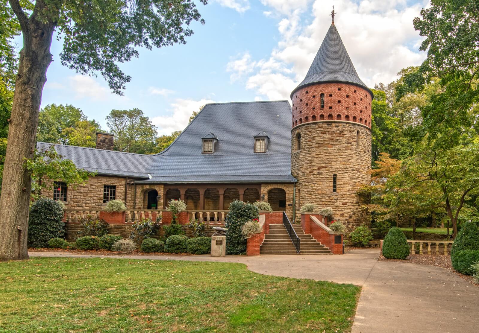 A stone building with a conical-roofed round tower, arched entryway, and surrounding trees under a partly cloudy sky.