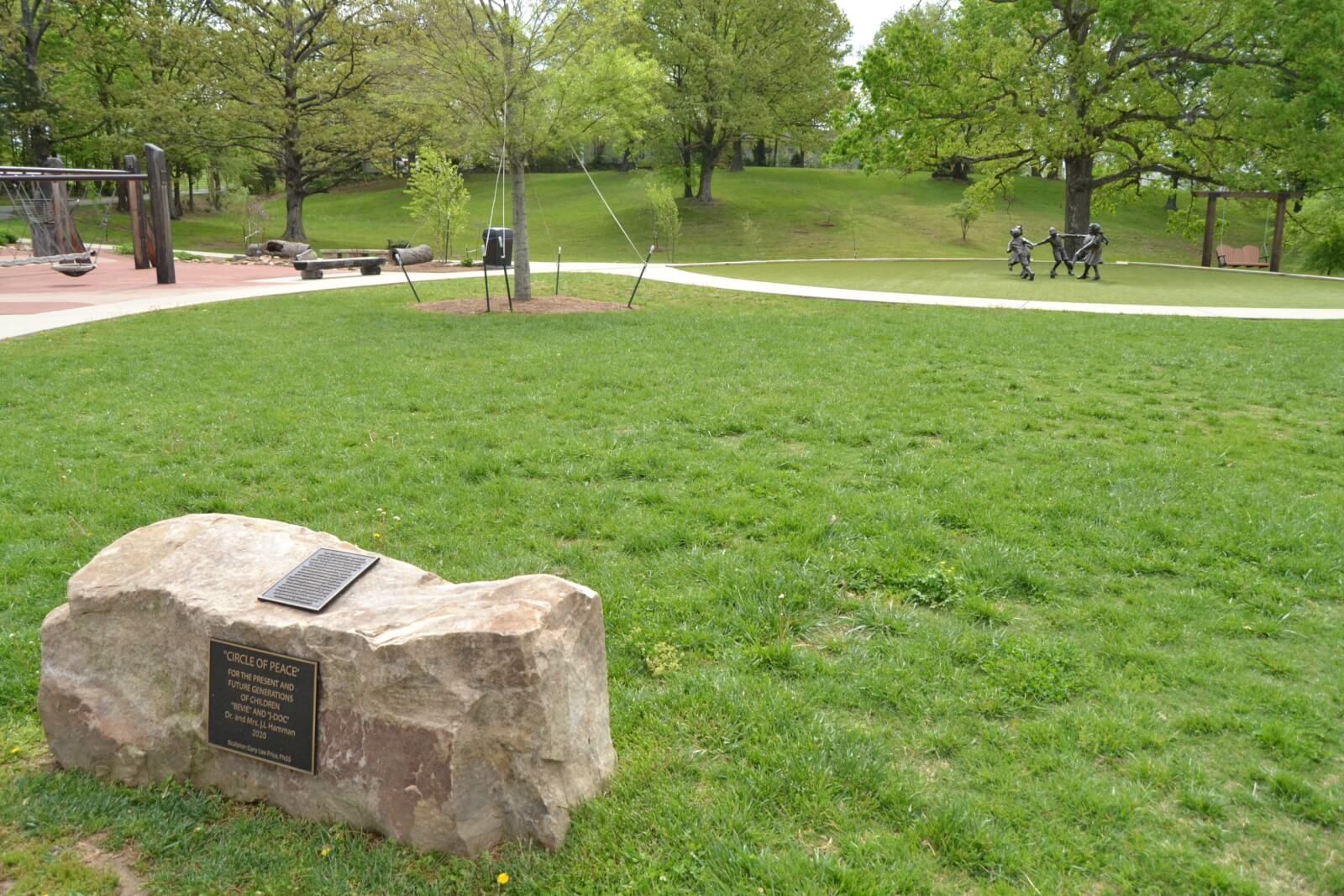 A large rock with a plaque sits on a grassy field near a paved path in a Western Kentucky park, where small towns offer big experiences among trees, a playground, and a metal sculpture in the background.