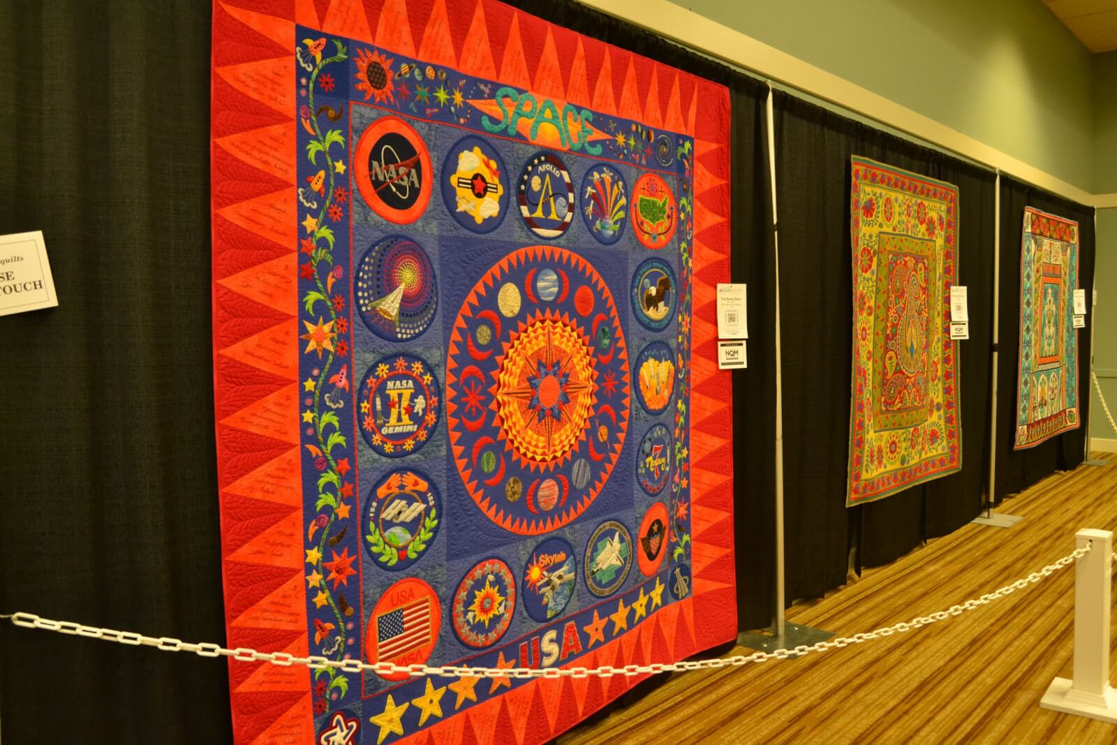 A colorful quilt featuring NASA and space-themed designs hangs on display alongside two other decorative quilts, highlighting the big experiences found in small towns like those in Western Kentucky.