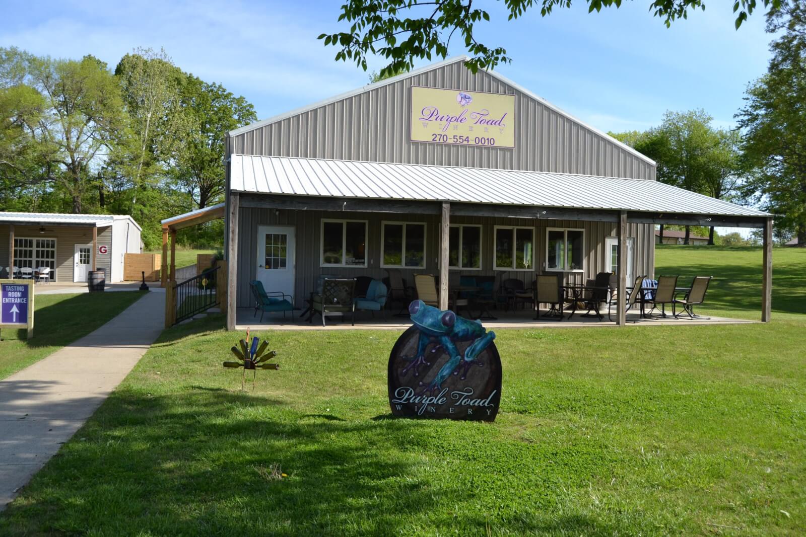 A metal building with a covered patio and outdoor seating sits on a grassy lawn in Western Kentucky, featuring a "Purple Toad" sign, trees, and another small town building in the background.