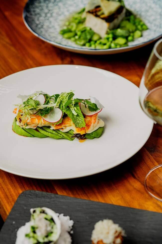 Where to Get Mother's Day Brunch in Memphis - 2 A plated dish featuring sliced avocado, shrimp, radish, and fresh herbs is perfect for a Mother's Day brunch in Memphis, with a glass of white wine and another entrée visible in the background.