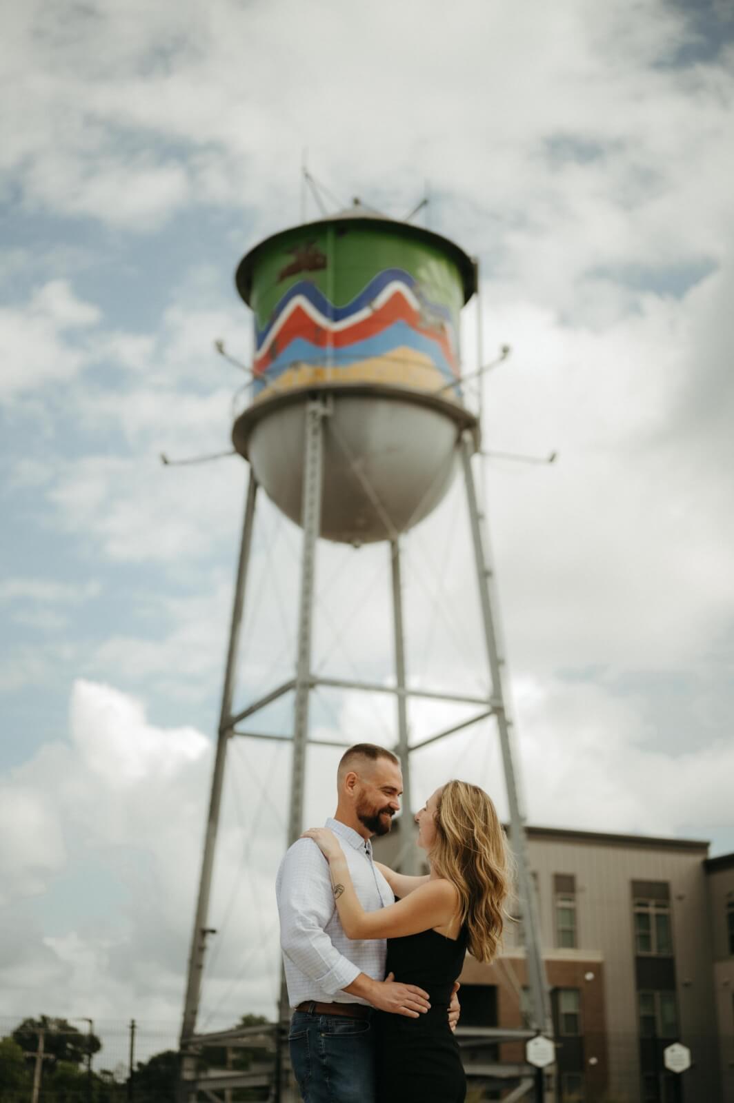 A couple stands closely together, looking at each other, in front of a tall, colorful water tower featuring vibrant Memphis street art on a partly cloudy day.