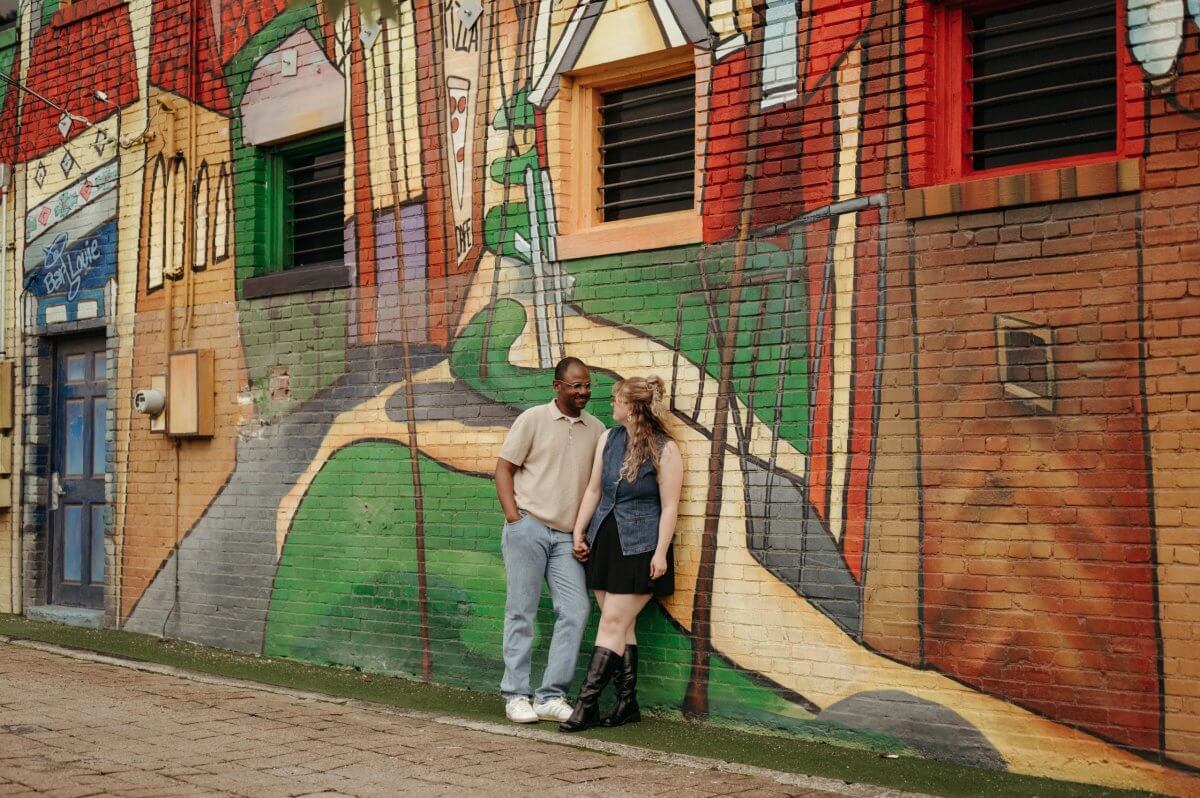 A man and woman stand holding hands in front of a colorful Memphis Murals masterpiece painted on a brick wall in an urban area.
