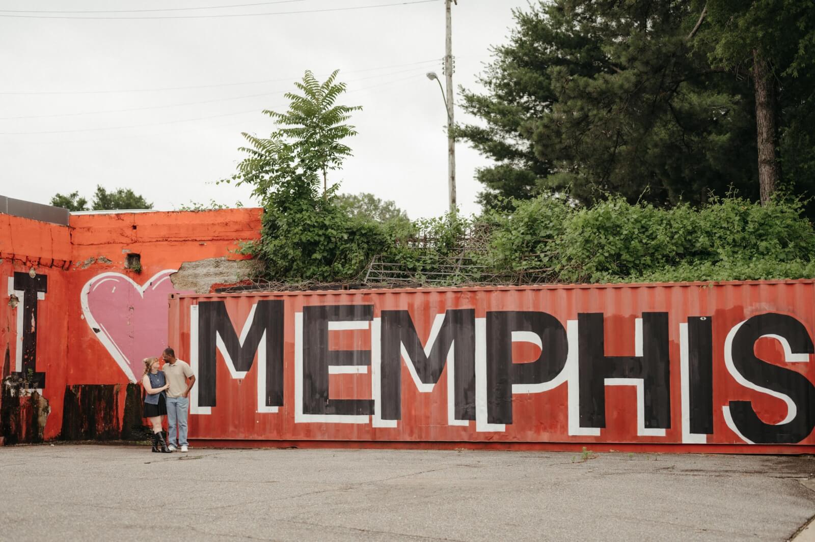A couple stands with a dog in front of a red "I ♥ MEMPHIS" mural, enjoying Memphis street art with greenery visible above the wall.