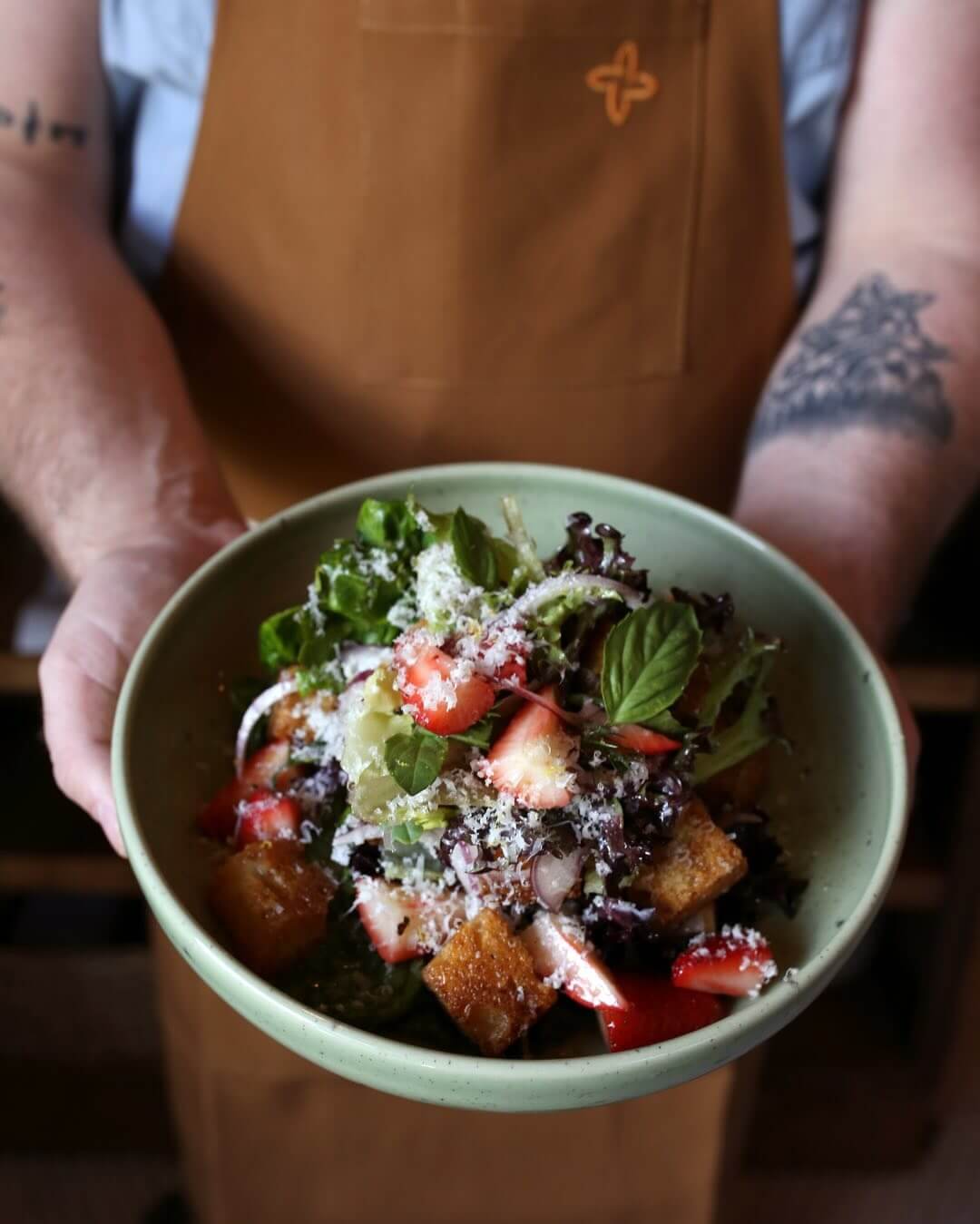 A person wearing a brown apron holds a green bowl filled with leafy greens, strawberries, croutons, onions, and grated cheese.