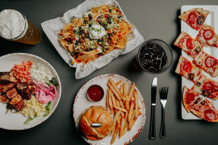A burger with fries, a salad with grilled meat, nachos with sour cream, pepperoni pizza slices, soda, and beer are arranged on a table—perfect for sampling the flavors at new Memphis restaurants.