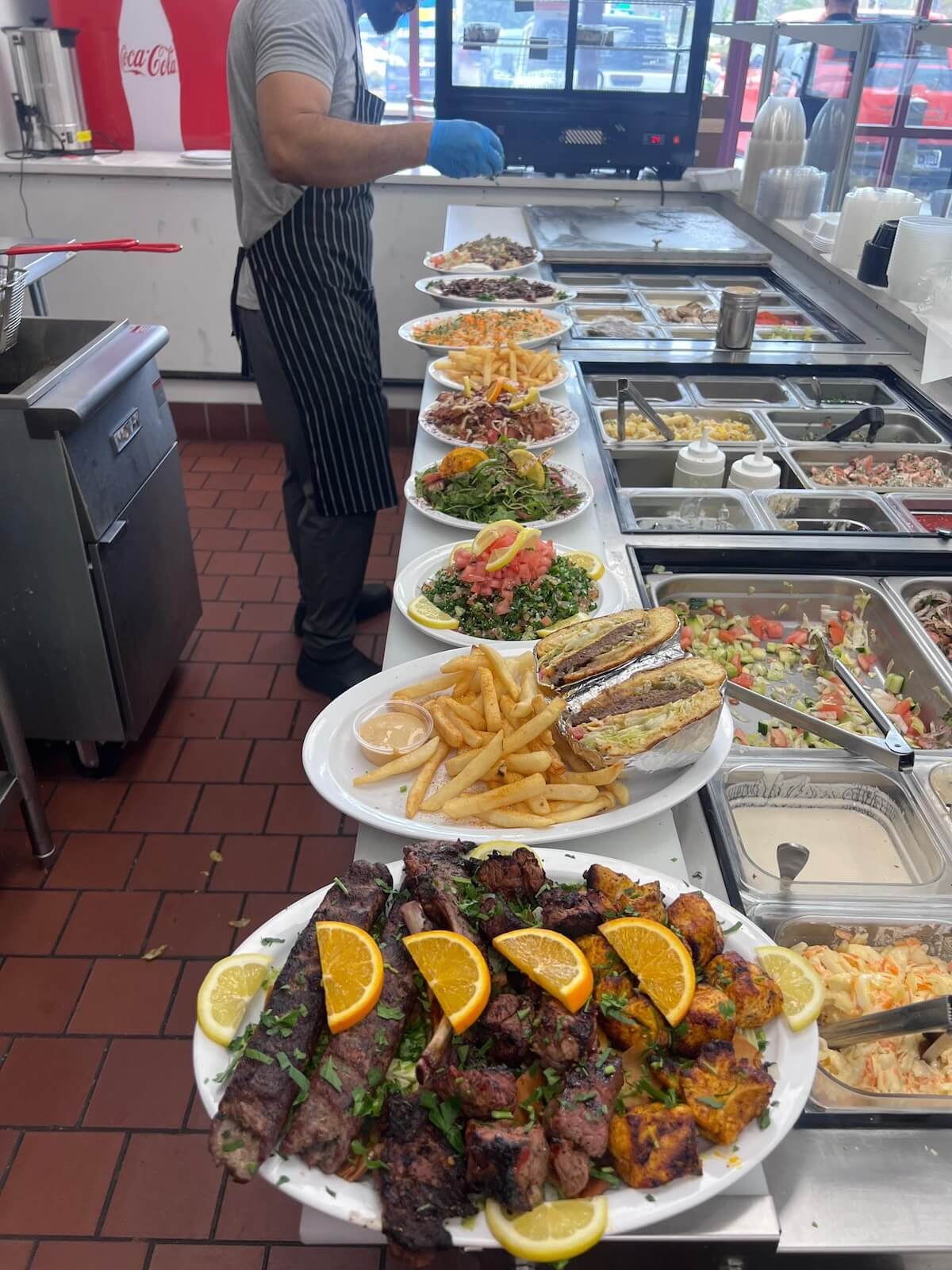 Several plates of Mediterranean-style dishes, including kebabs, fries, salad, and sandwiches, are lined up on a counter in a new Memphis restaurant kitchen with a worker preparing food.