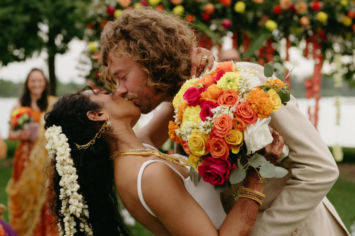 A couple kisses as they embrace outdoors at the Swamy-Collins Wedding; the woman holds a colorful bouquet of roses and marigolds, with wedding guests visible in the background.