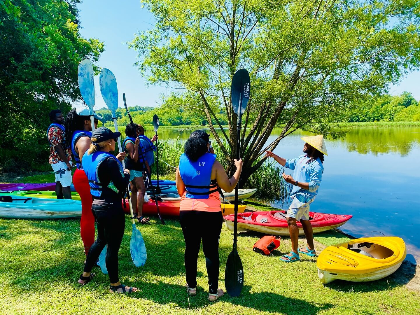 A group of people wearing life jackets stand by kayaks on grass near a lake, listening to an instructor from Capital City Kayaks 