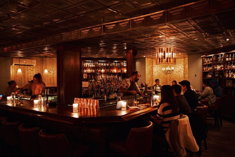 Dimly lit bar in The Gulch with patrons at the counter and tables, bartender serving drinks, shelves of bottles behind the bar, and candles providing warm ambient light.