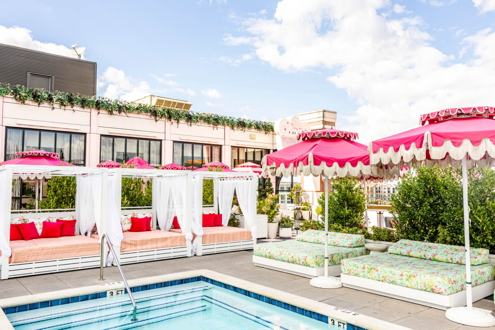 Chic outdoor hotel pools area featuring striped cabanas, patterned lounge beds, pink umbrellas, and lush greenery on a rooftop with city views under a partly cloudy sky.