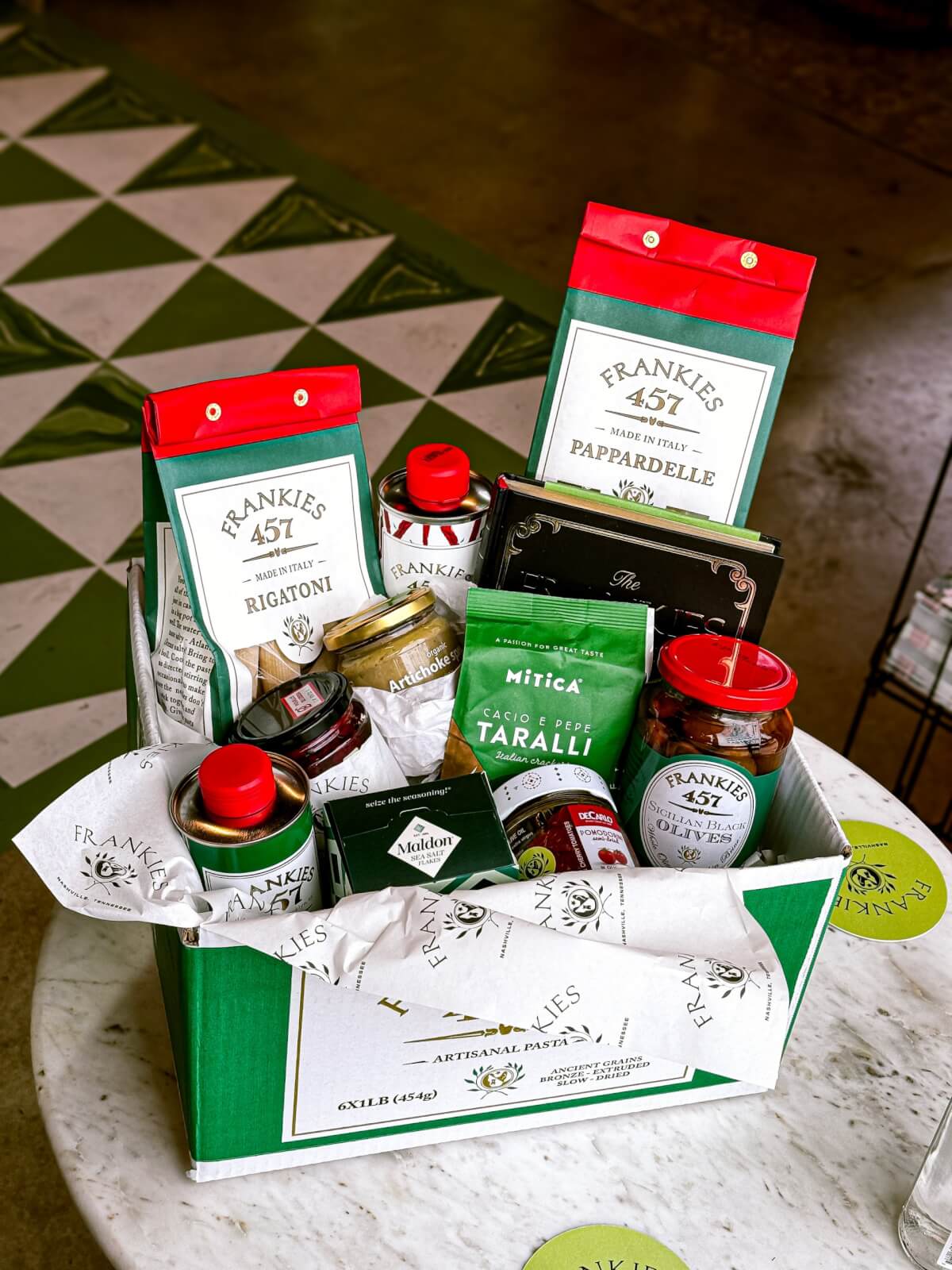 A green gift box on a marble table filled with Italian food items—perfect for June gifting in Nashville—includes pasta, olive oil, sauce jars, taralli, and chocolate, with patterned floor tiles in the background. A true Southern FINDS treat.