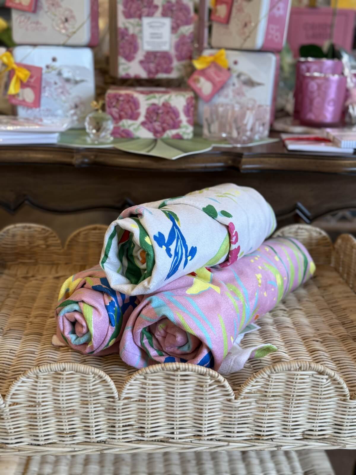 Three rolled, floral-patterned towels are displayed on a woven wicker tray, with boxed floral-themed products in the background—perfect for your June FINDS in Nashville.