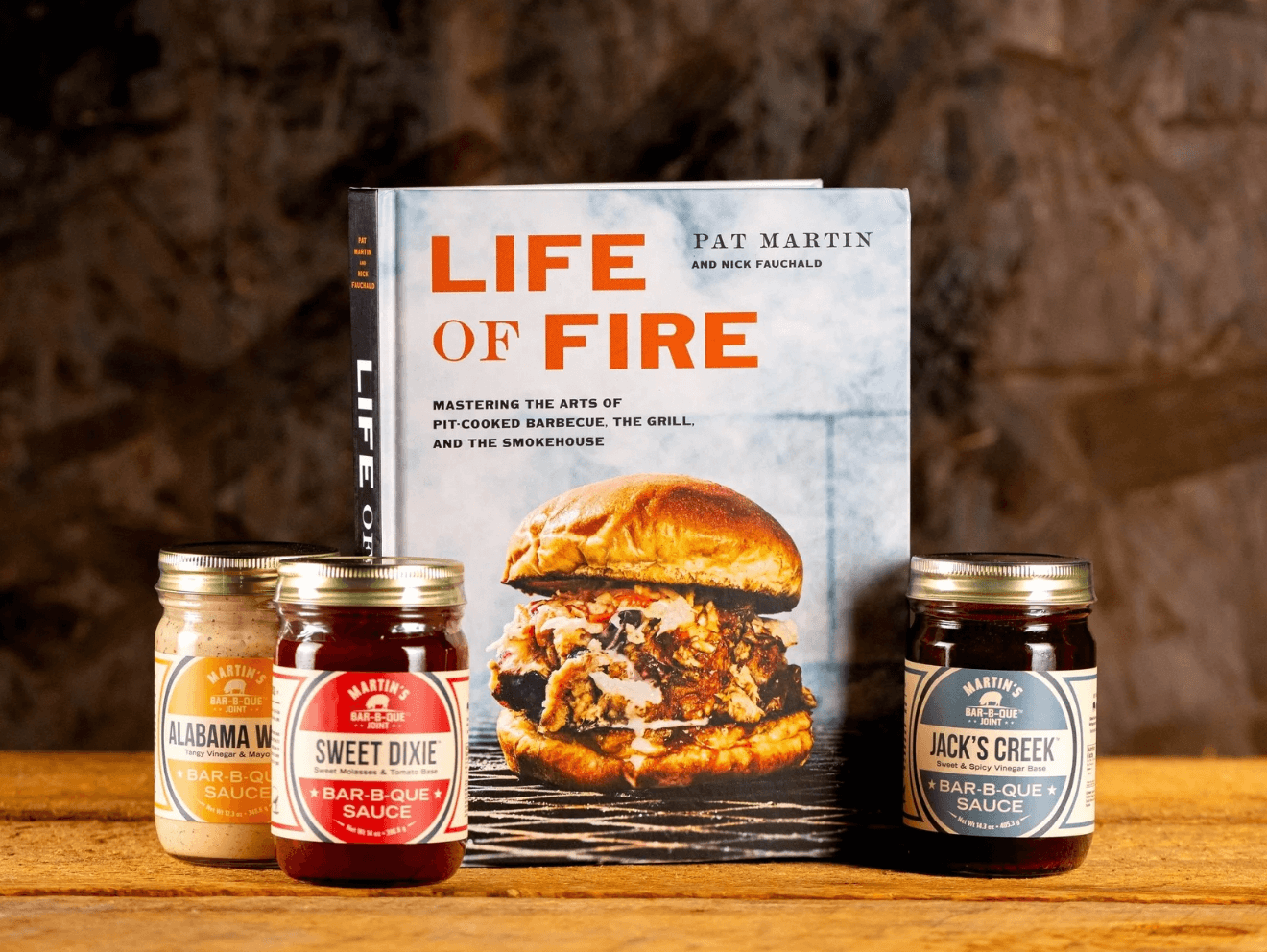 A cookbook titled "Life of Fire" stands behind three jars of barbecue sauces—Alabama White, Sweet Dixie, and Jack’s Creek—on a wooden surface, showcasing delicious Nashville FINDS perfect for June grilling.