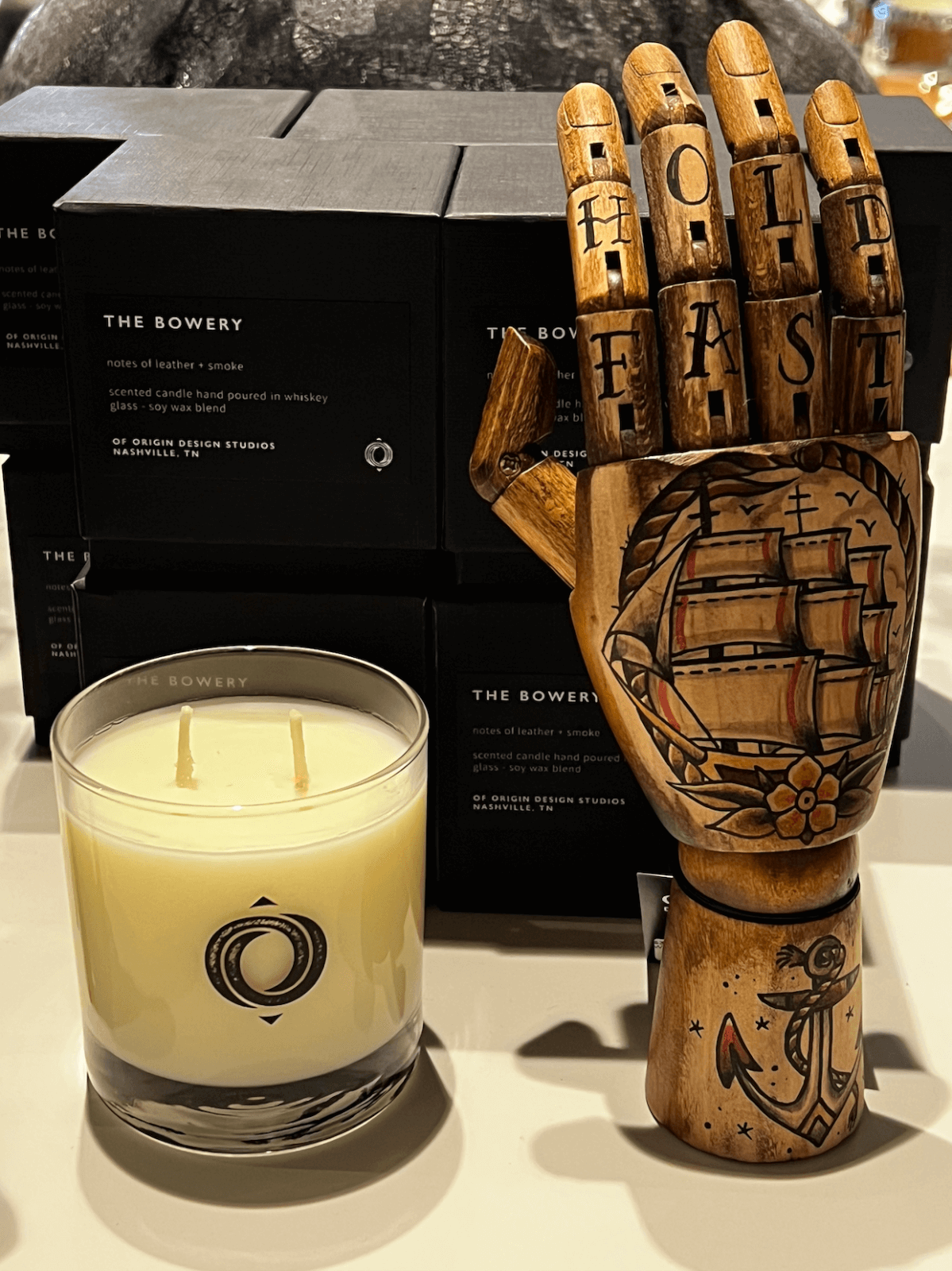 A candle and a wooden hand sculpture with "HOLD FAST" and a ship tattoo are displayed in front of stacked black boxes labeled "The Bowery"—a standout June FINDS discovery in Nashville.