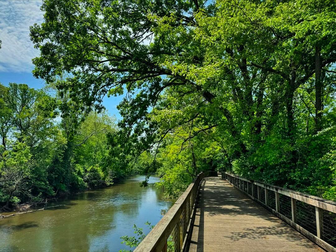 A wooden boardwalk runs alongside a calm river, surrounded by lush green trees under a partly cloudy sky—one of the scenic greenways perfect for Nashville running routes.