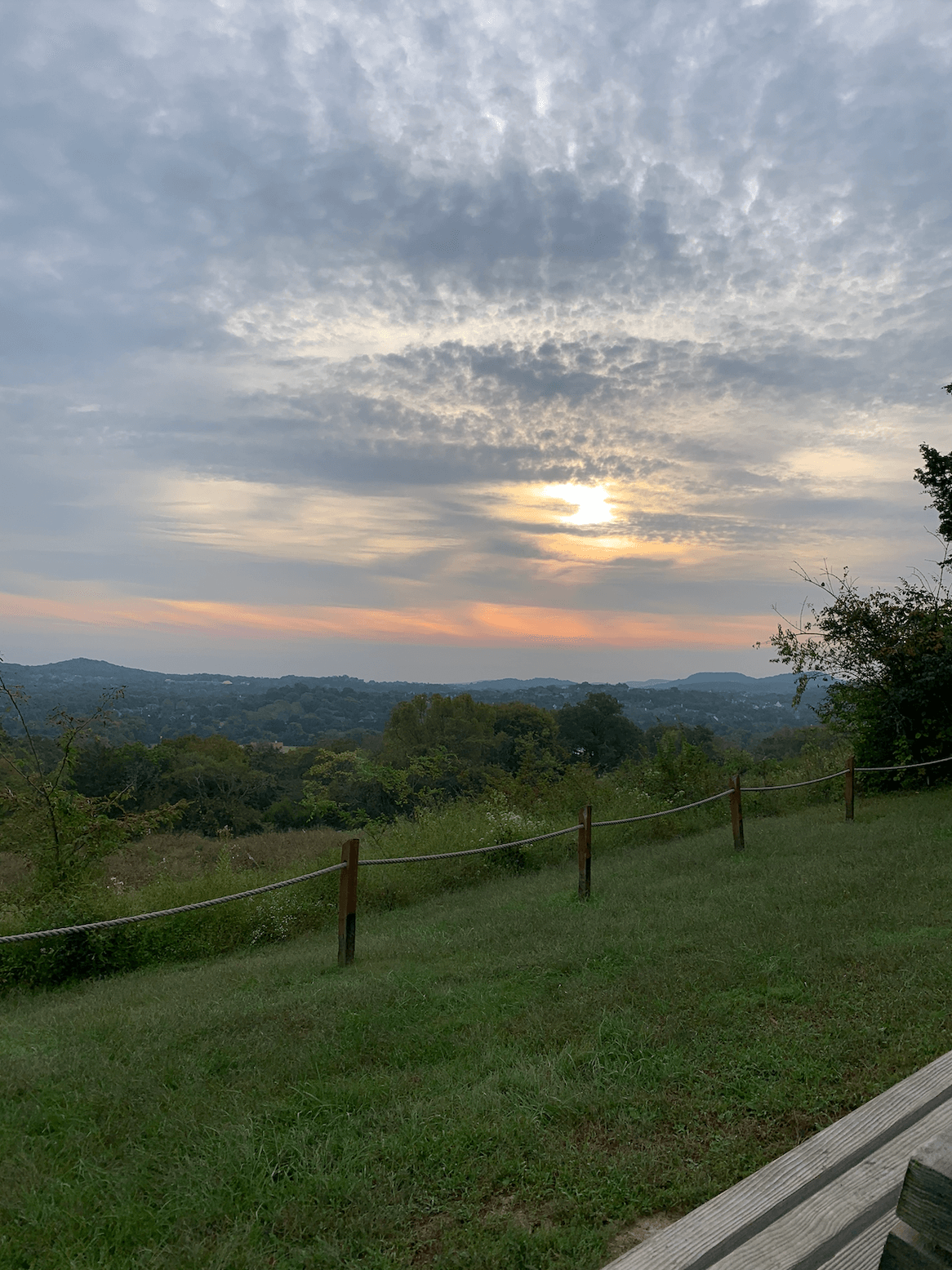 A cloudy sky at sunset over rolling hills and trees, with a grassy area and wooden fence in the foreground—perfect scenery often found along Nashville running routes and scenic greenways.