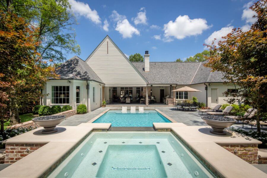 A modern house with a backyard featuring a swanky pool design, hot tub in the foreground, lounge chairs, and landscaped trees under a blue sky.