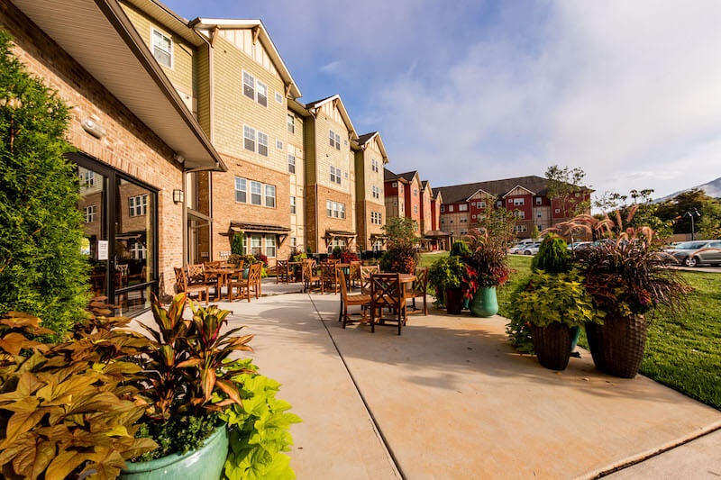 Dedicated to Hurricane Helene Recovery, This NC Architect is Donating Her Time - 2 A patio with wooden tables and chairs is set outside a multi-story brick building, thoughtfully designed by an architect, surrounded by potted plants and landscaped greenery under a partly cloudy sky.
