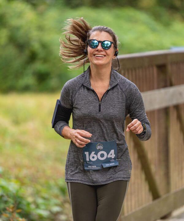 Dedicated to Hurricane Helene Recovery, This NC Architect is Donating Her Time - 1 A woman wearing sunglasses, a gray long-sleeve shirt, and a race bib numbered 1604 runs outdoors on a path beside a wooden fence.