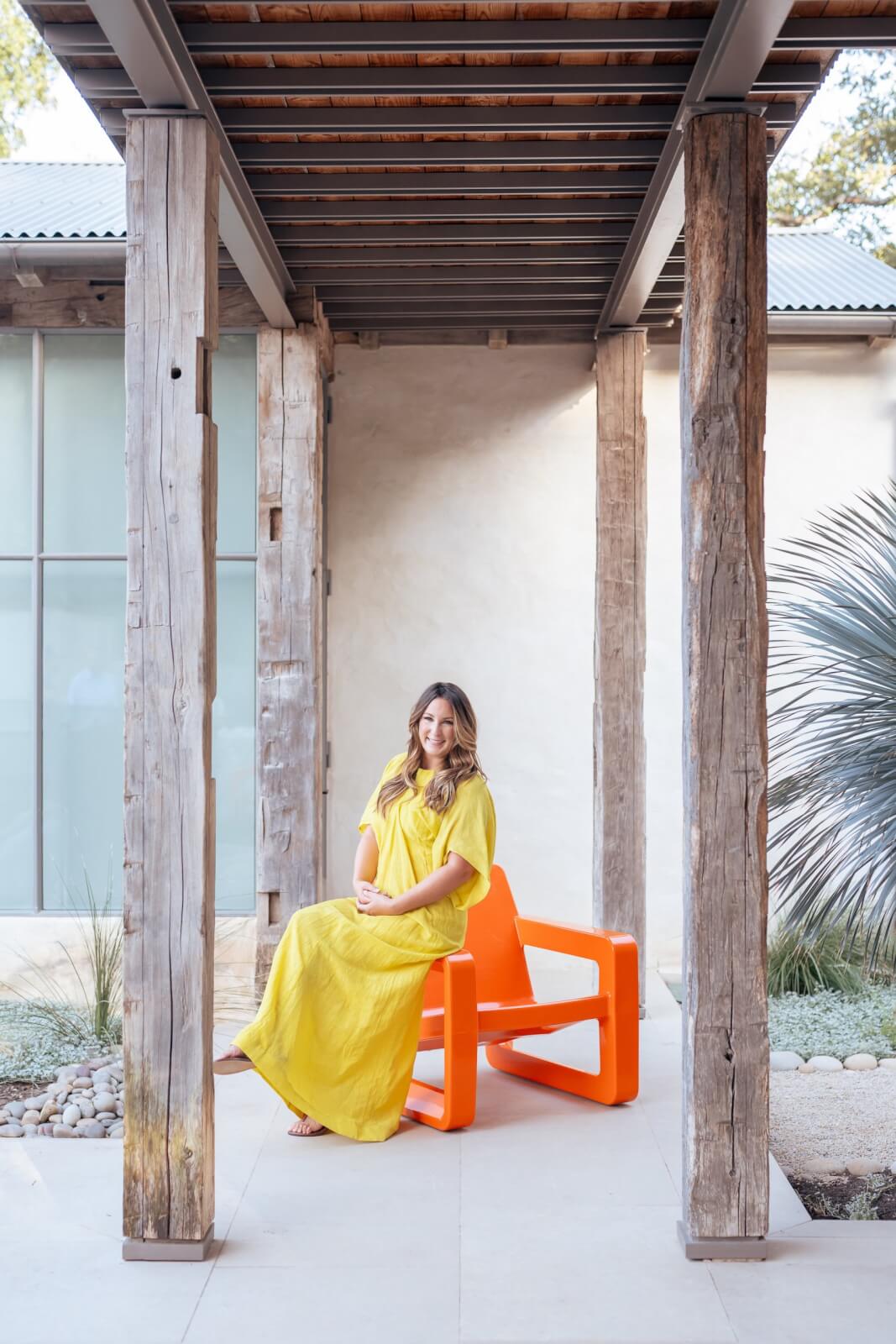 Sara Kenton, in a yellow dress, sits on an orange Tupelo Goods chair under a wooden pergola, with a neutral-toned building and desert landscaping in the background.