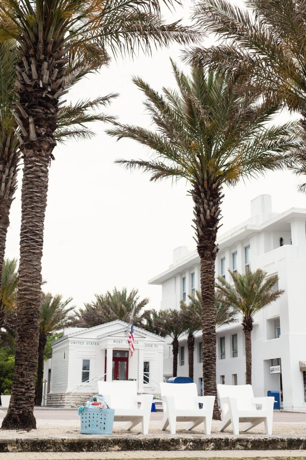 A white post office building with a US flag stands behind palm trees and white outdoor chairs by Tupelo Goods; a laundry basket sits in the foreground, capturing the relaxed charm of Sara Kenton's design.