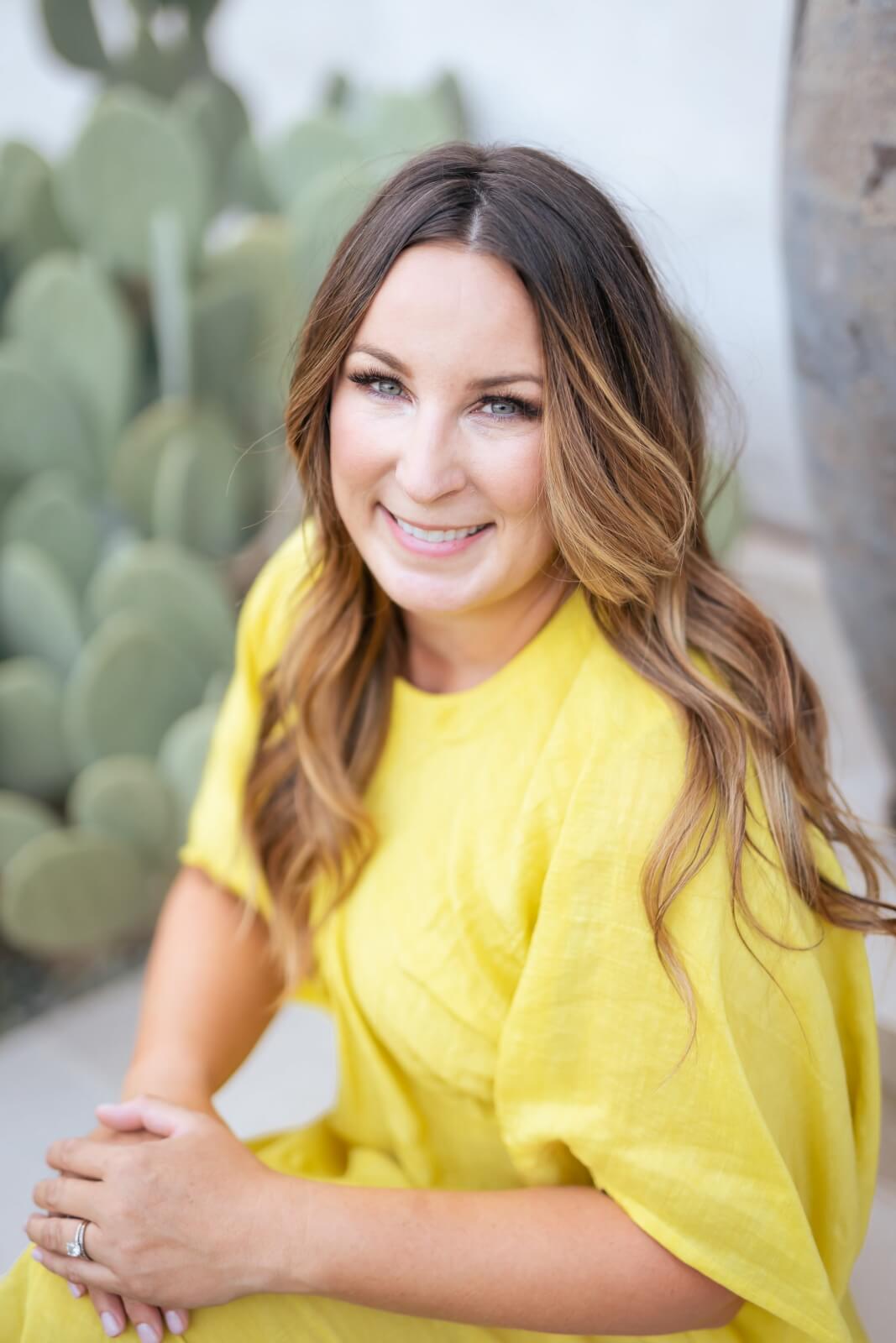 Sara Kenton, with long brown hair, sits outdoors in a bright yellow dress, smiling at the camera in front of green cacti.
