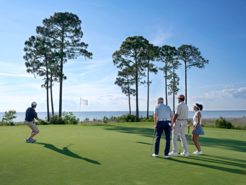 5 Reasons South Walton is Becoming the Premier Destination for Corporate Events and Meetings - 7 Four people stand on a golf green near the hole, with one person celebrating. Set in South Walton, this picturesque event venue features tall trees and water beyond the course under a blue sky.