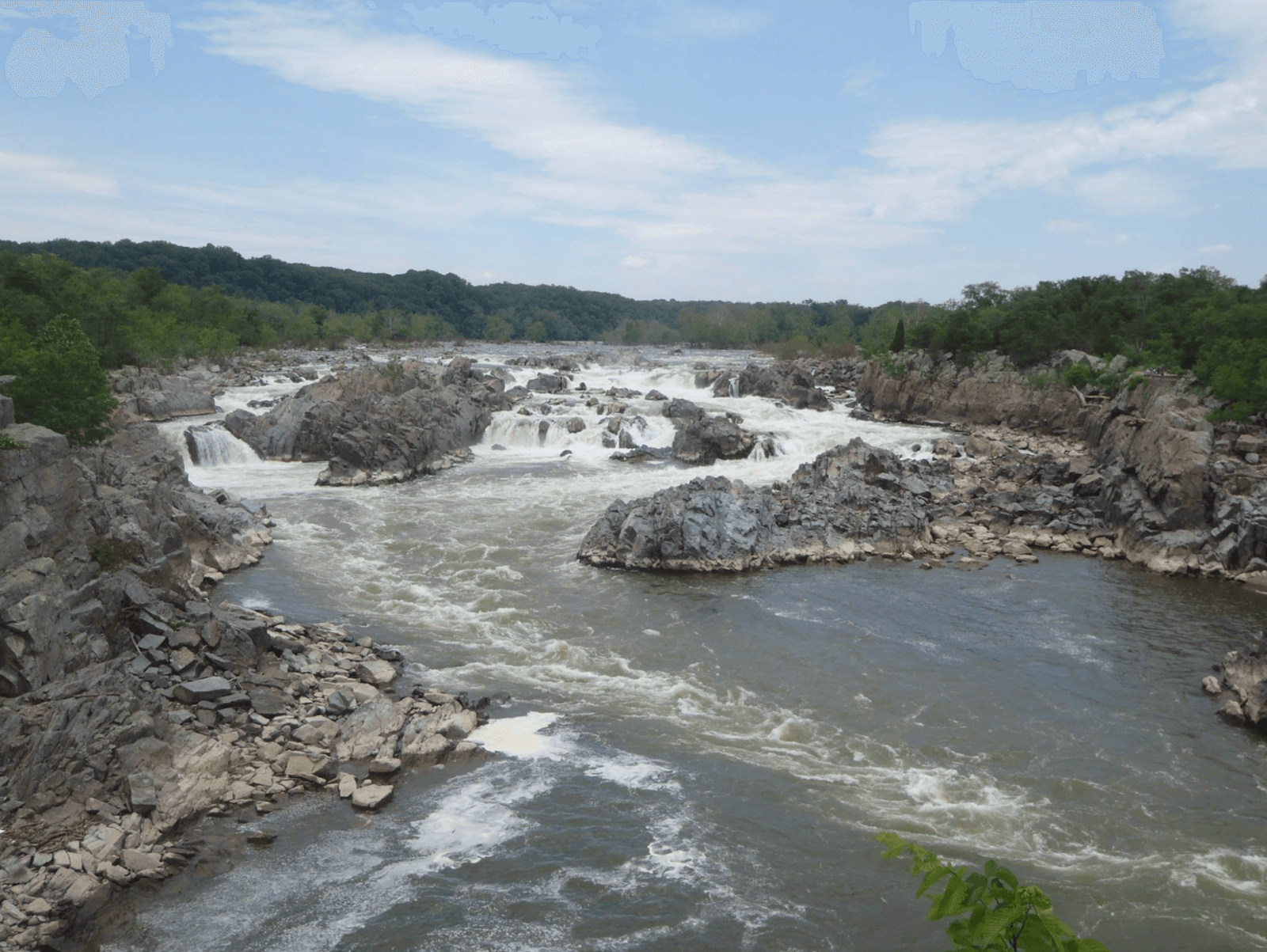 A wide river flows over rocky ledges and small waterfalls, perfect for waterfall hikes in Virginia, surrounded by rugged rocks and green trees under a partly cloudy sky.