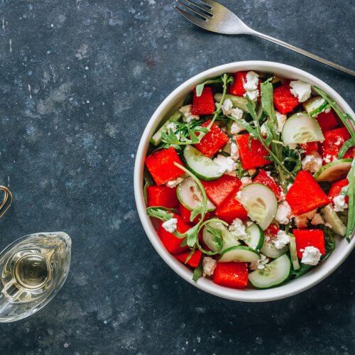A bowl of salad featuring watermelon, cucumber, arugula, and feta cheese sits on a dark countertop—perfect for those seeking refreshing watermelon recipes. A fork, oil bottle, and whisk rest nearby.