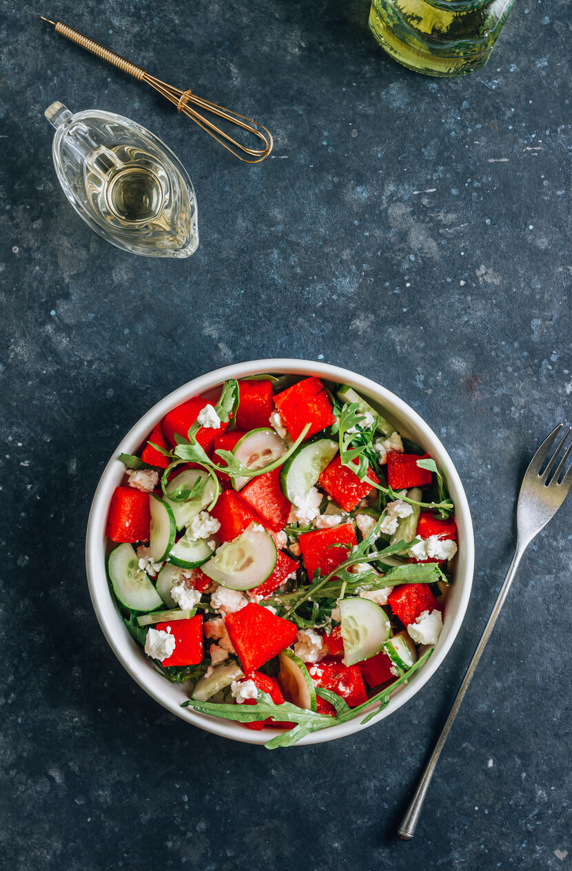 A bowl of salad with watermelon cubes, cucumber slices, arugula, and feta cheese sits on a dark surface next to a fork, a small glass pitcher, and a bottle of oil—perfect for easy watermelon recipes.