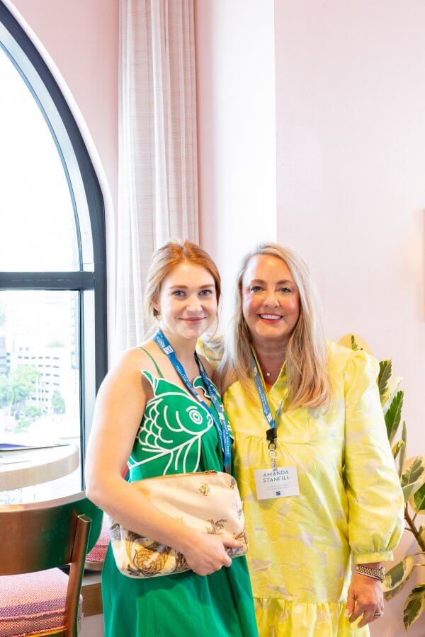 Two women standing indoors near a window, both wearing name badges and brightly colored clothing, posing and smiling for the camera.