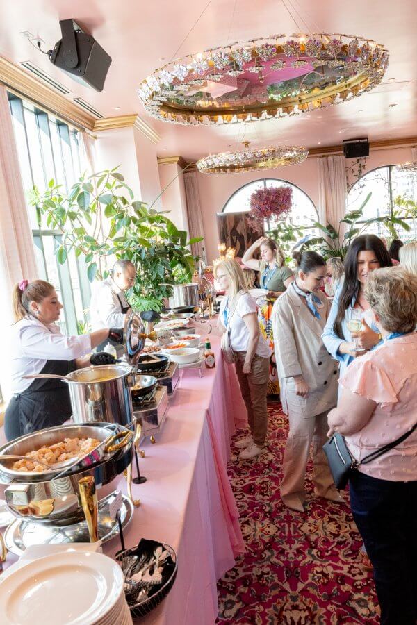 People converse and serve themselves food from a buffet table in a bright, plant-filled room with pink decor and ornate lighting.