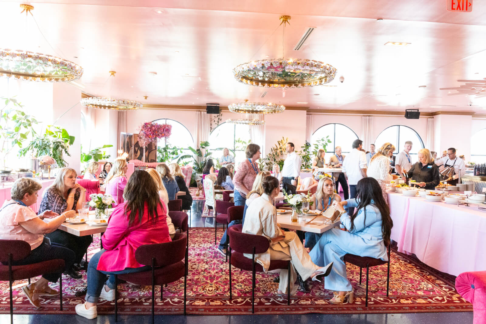 5 Reasons South Walton is Becoming the Premier Destination for Corporate Events and Meetings - 13 People seated and standing in a brightly lit, pink-themed dining room, engaging in conversation and eating at an event with buffet tables in the background.