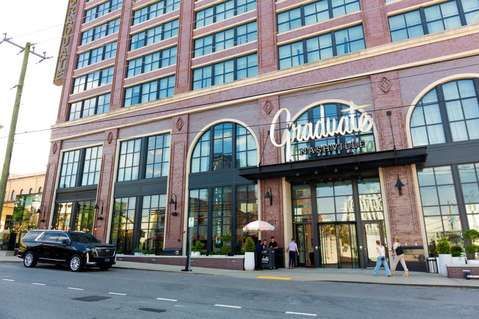 A brick building with large windows and a sign reading "Graduate Nashville" above the entrance; a black SUV is parked at the curb and several people stand by the door.