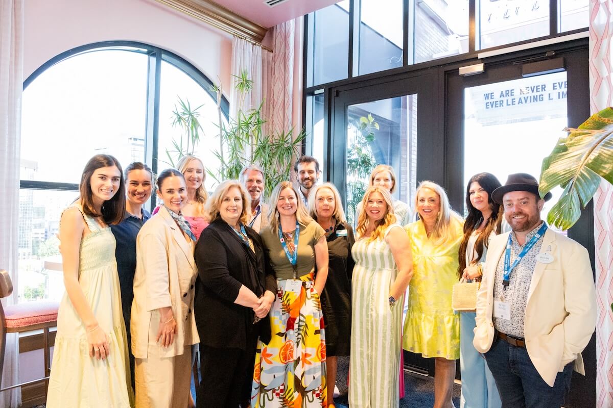 A group of thirteen adults pose together indoors near large windows, many wearing badges and business-casual attire, with natural light filling the room.